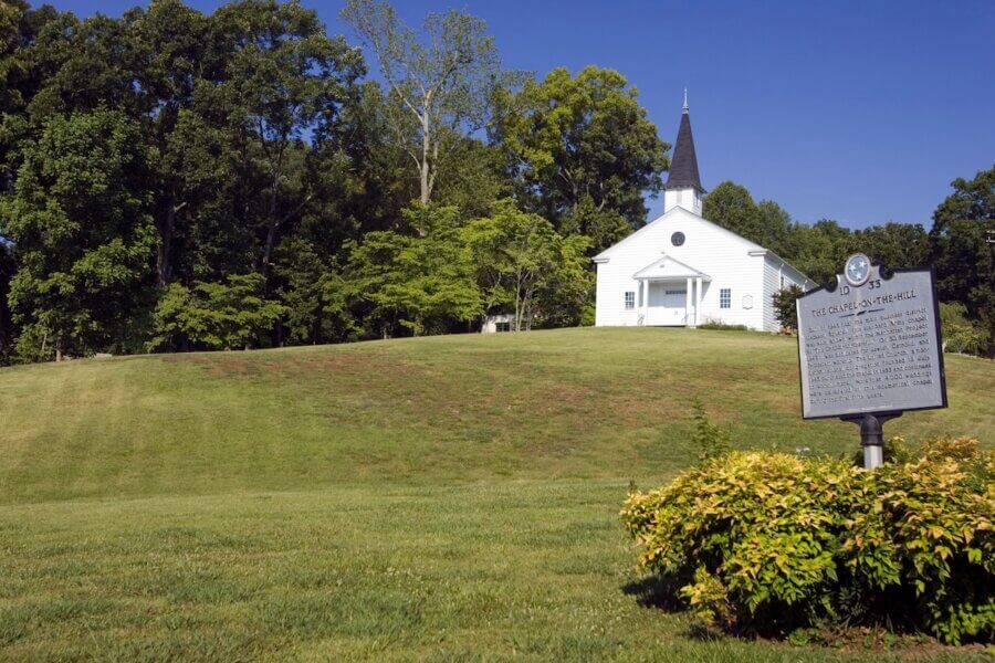 Living History: 7 Tennessee Towns Where the Past Comes Alive - 11 A small white church with a steeple sits on a grassy hill, surrounded by trees — a charming scene often found in historic Tennessee towns. A historical marker sign is in the foreground near bushes.