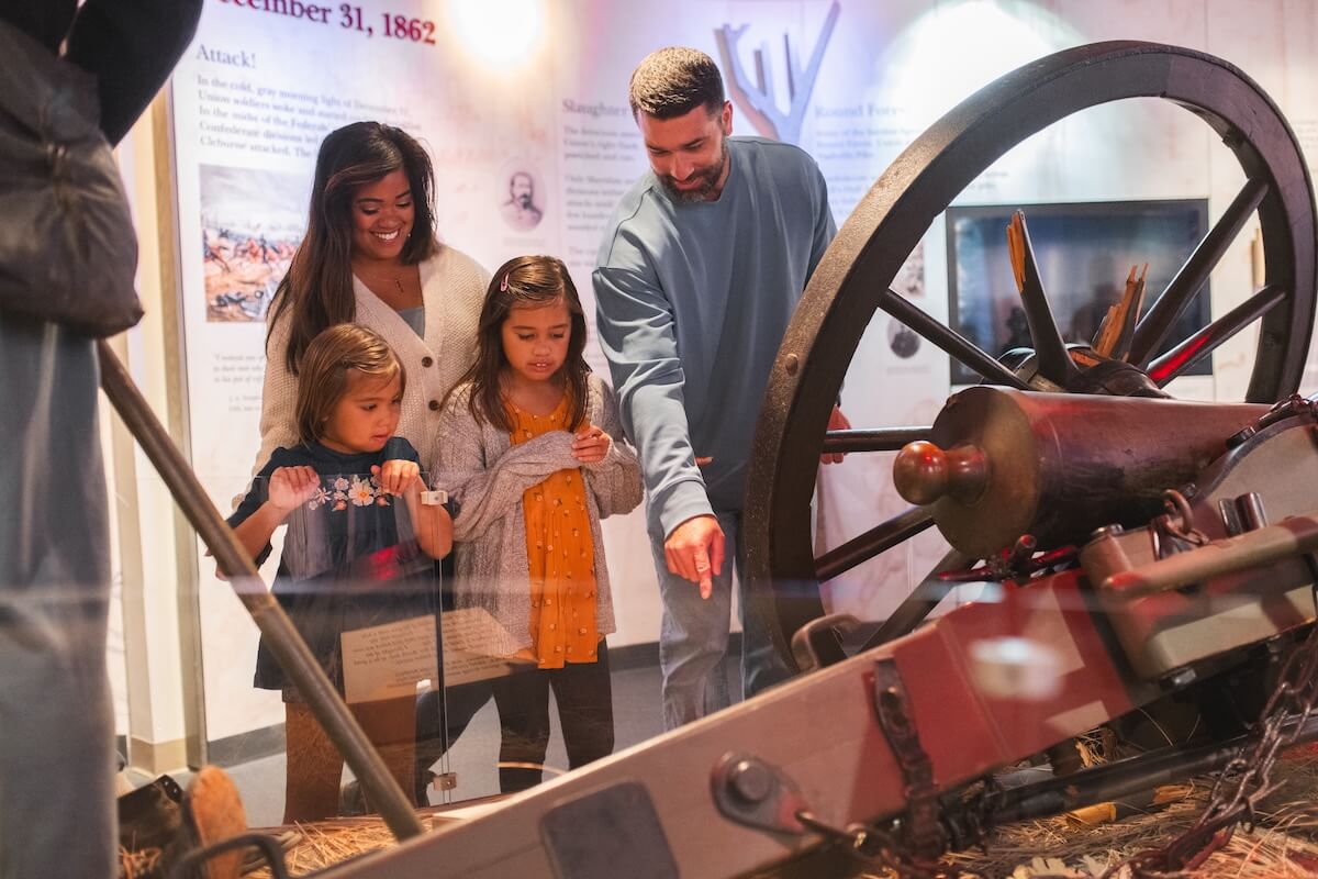 A family of four observes and discusses a historical cannon and artifacts in a museum exhibit, immersing themselves in the living history found in Tennessee towns.