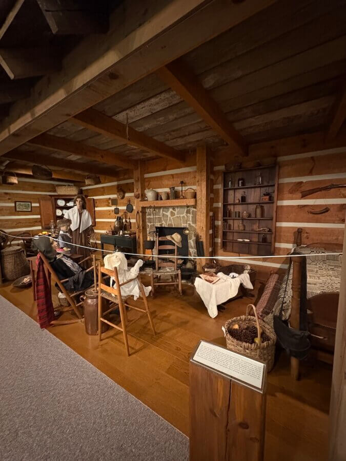 Living History: 7 Tennessee Towns Where the Past Comes Alive - 8 A living history log cabin interior display with a stone fireplace, period furniture, household items, mannequins in old-fashioned clothing, and various tools and baskets — evoking the charm of early Tennessee towns.