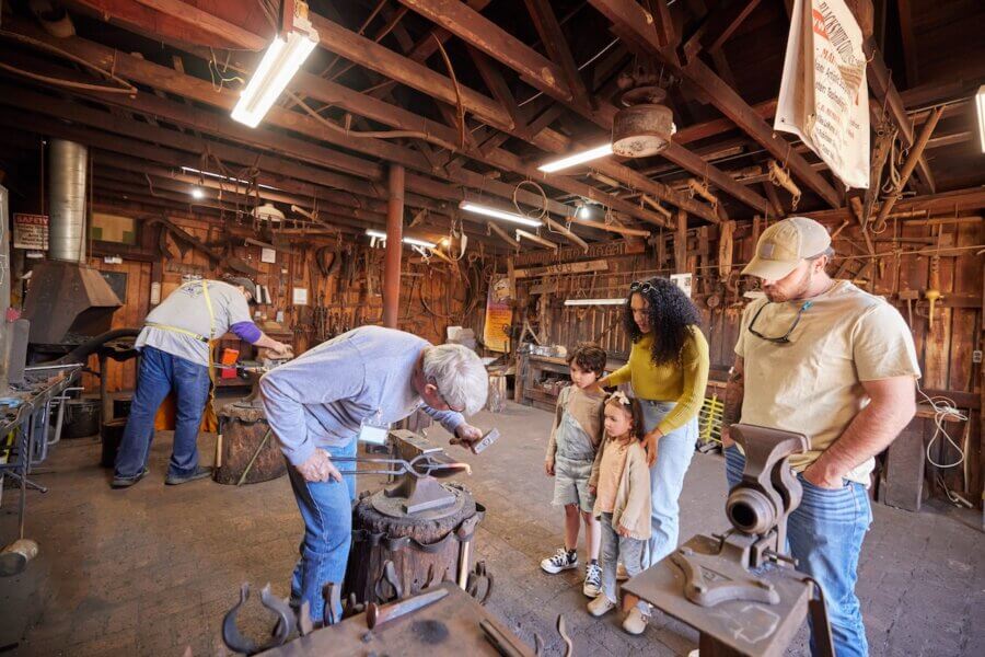 Living History: 7 Tennessee Towns Where the Past Comes Alive - 2 A blacksmith demonstrates metalworking to a family with two children inside a workshop, bringing living history to life, while another person works at a separate station in the background.