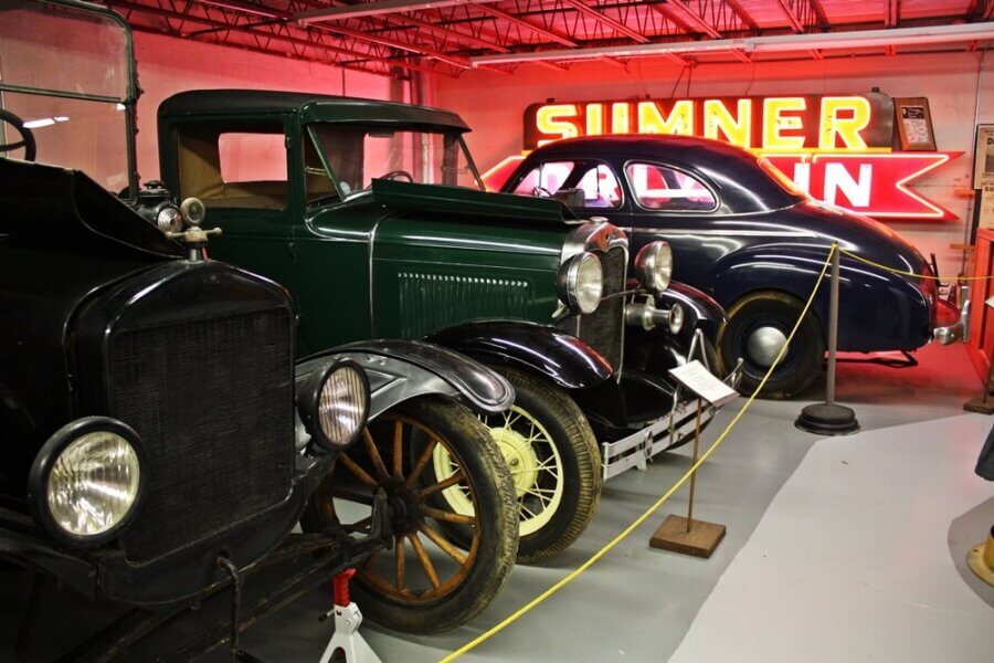Living History: 7 Tennessee Towns Where the Past Comes Alive - 6 Three vintage cars are displayed in a museum, capturing the spirit of Tennessee towns, with a large red "SUMNER" neon sign glowing in the background and a yellow rope barrier in front.