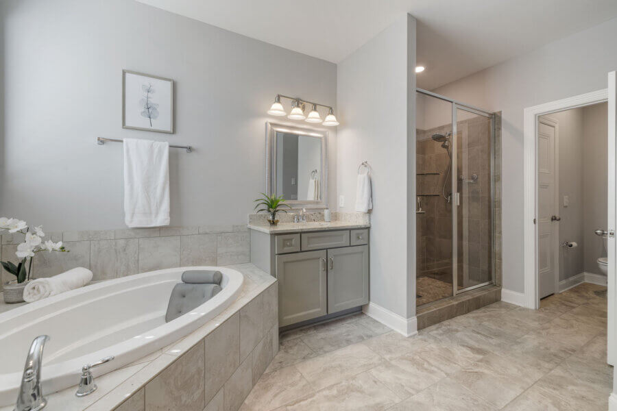Just Listed! This Nolensville Home is Ready for Summer Entertaining - 11 Modern bathroom in a Nolensville Home with a large bathtub, gray vanity with mirror, glass-enclosed shower, and tiled floor; minimal decor and neutral color scheme—perfect for relaxing after summer entertaining.
