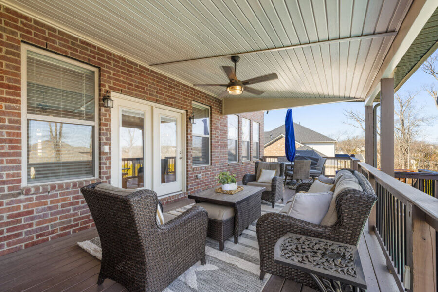 Just Listed! This Nolensville Home is Ready for Summer Entertaining - 8 Covered porch with wicker furniture, ceiling fan, rug, and potted plant, attached to a brick Nolensville Home with windows and glass doors—perfect for summer entertaining overlooking a charming residential neighborhood. Just listed!.