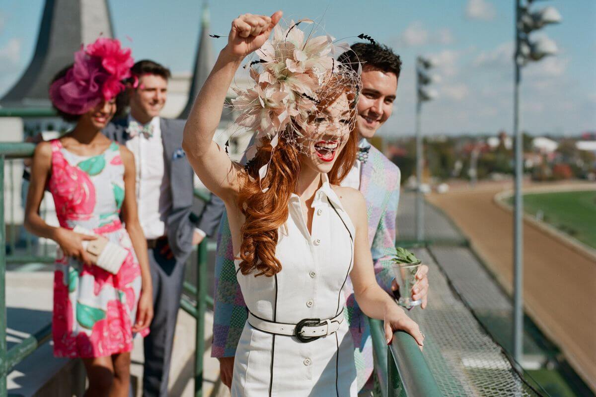 A woman in a white dress and floral hat cheers at the Thoroughbred horse races, accompanied by three well-dressed people, with the racetrack and stands visible in the background—a perfect travel experience.