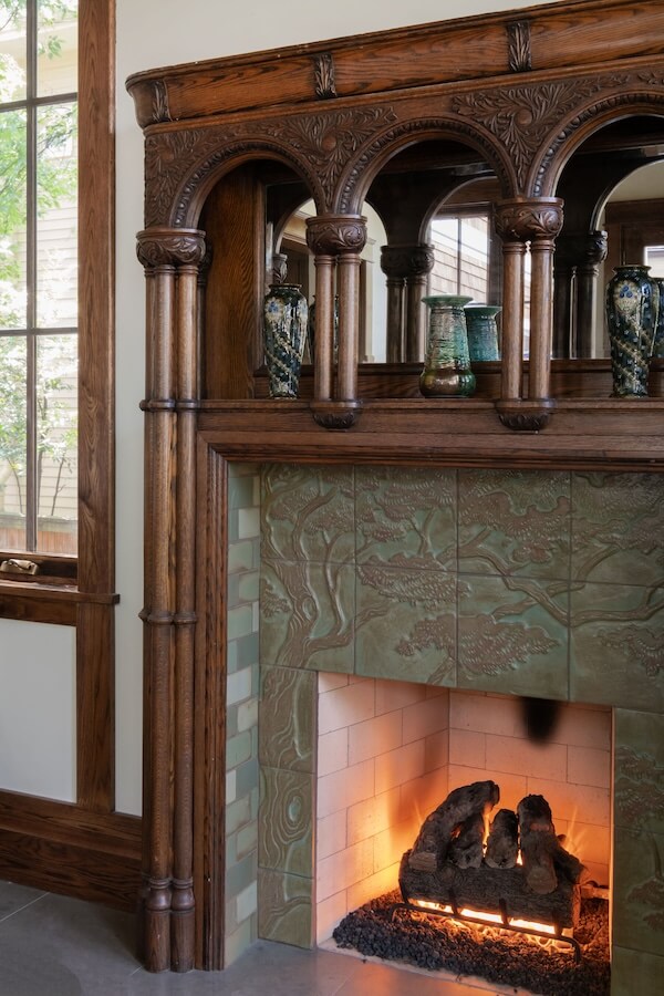 7 Tile Trends Interior Designers Are Loving Right Now - 5 Ornate wooden mantel with decorative columns and vases above a lit fireplace featuring green tile trends and a relief tree design, next to a window.