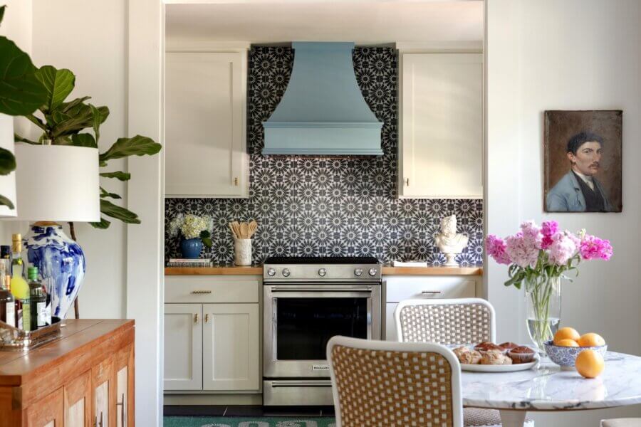 7 Tile Trends Interior Designers Are Loving Right Now - 4 A kitchen features white cabinets, a stainless steel stove, a blue range hood, and a patterned tile backsplash that highlights the latest tile trends. A small dining table with flowers and pastries sits nearby, with a portrait hanging on the right wall.