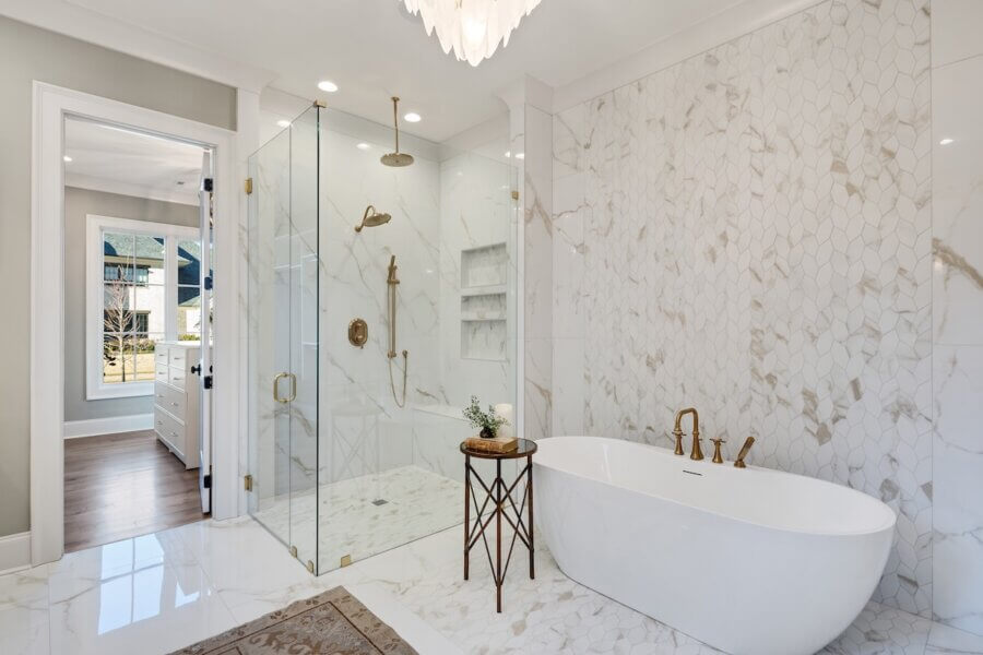 7 Tile Trends Interior Designers Are Loving Right Now - 8 Modern bathroom with glass-enclosed shower, freestanding bathtub, marble tile walls and floors showcasing the latest tile trends, gold fixtures, and a small round side table.