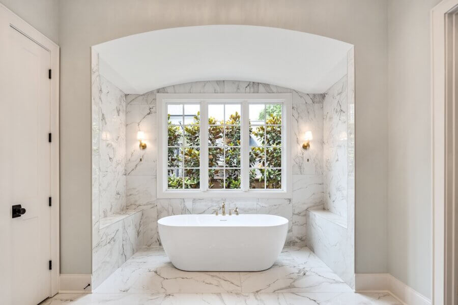 7 Tile Trends Interior Designers Are Loving Right Now - 13 Modern bathroom featuring a freestanding white bathtub, marble walls and floors reflecting the latest tile trends, a large window, and wall sconces on either side.