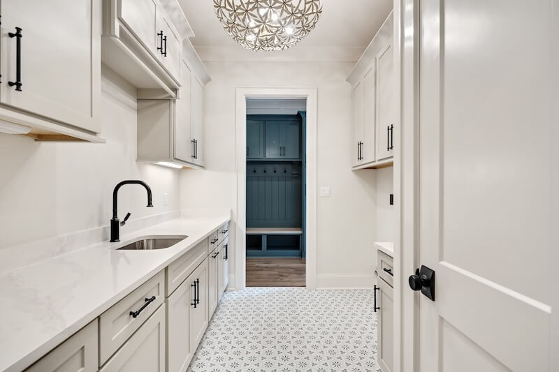 7 Tile Trends Interior Designers Are Loving Right Now - 19 Modern laundry room with white cabinets, black hardware, a sink, and a patterned tile floor inspired by the latest tile trends, plus a decorative light fixture, leading to a mudroom with built-in storage benches.