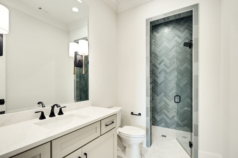 7 Tile Trends Interior Designers Are Loving Right Now - 17 Modern bathroom with a double vanity, white countertop, black fixtures, and a walk-in shower featuring green herringbone tile—highlighting current tile trends—with a glass door and a sleek toilet.