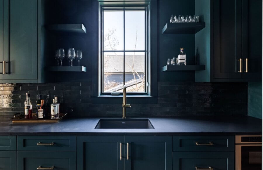 7 Tile Trends Interior Designers Are Loving Right Now - 2 A modern kitchen features teal cabinets, a brass faucet, glassware on shelves, liquor bottles on the counter, and a window above the sink with natural light highlighting the latest tile trends.