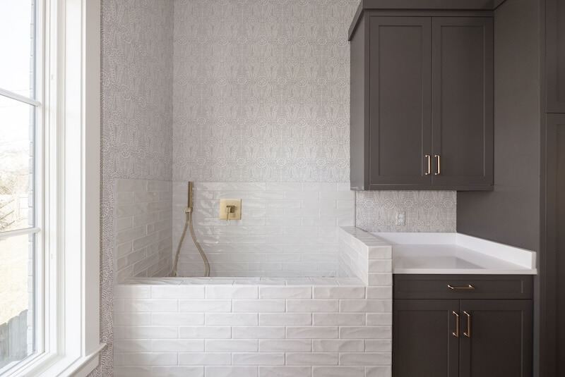 7 Tile Trends Interior Designers Are Loving Right Now - 23 Laundry room with a large utility sink, gray cabinets, and a white subway tile backsplash featuring the latest tile trends, plus a window providing natural light.