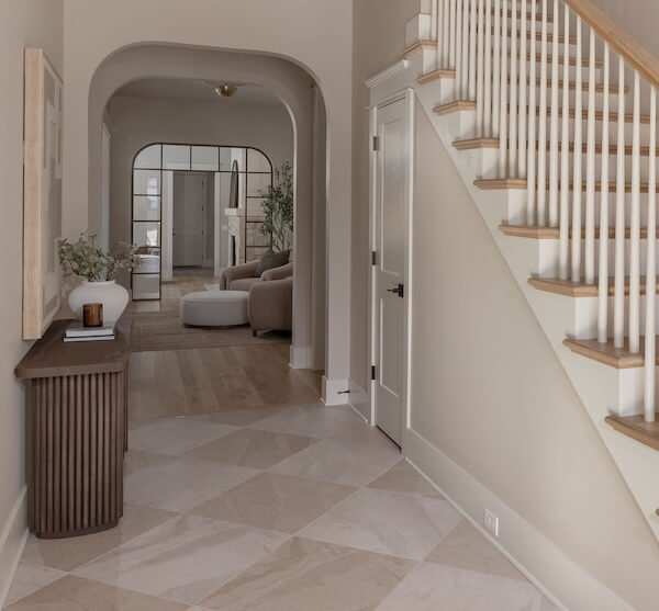 7 Tile Trends Interior Designers Are Loving Right Now - 14 A modern entryway with checkered tile flooring that showcases the latest tile trends, a wooden console table with decor, a staircase with white spindles, and a view into a living area with neutral furnishings.