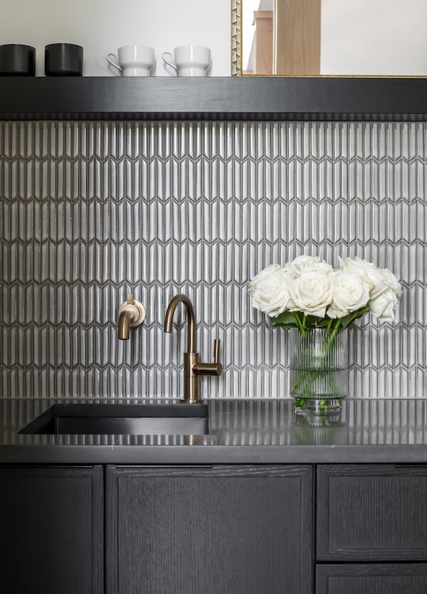 7 Tile Trends Interior Designers Are Loving Right Now - 25 A modern kitchen counter with a black sink, bronze faucet, tile trends showcased in the patterned backsplash, and a glass vase of white roses next to dark cabinetry.