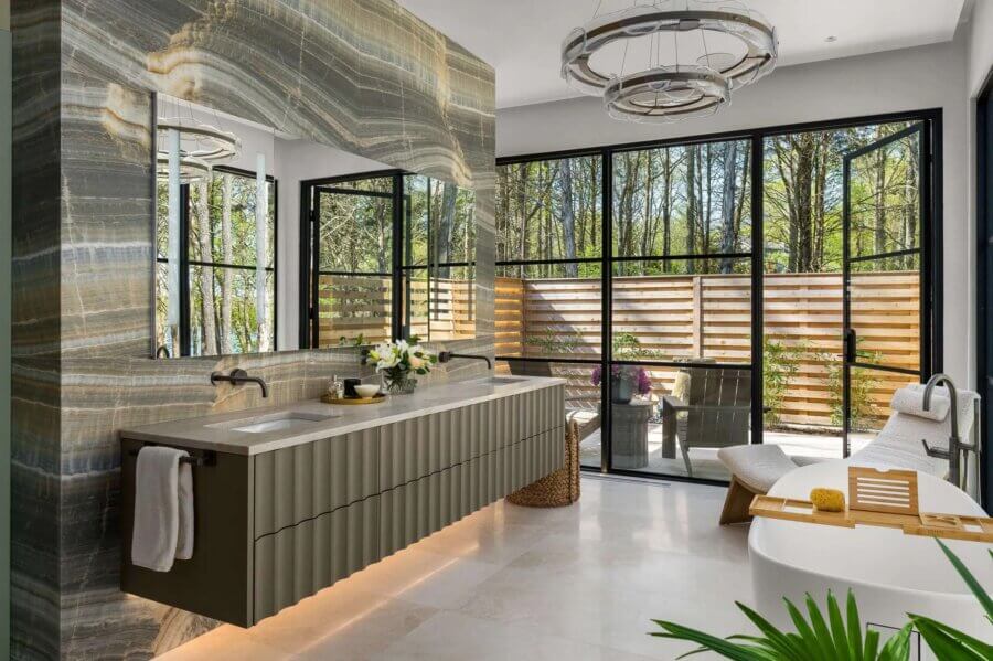 7 Tile Trends Interior Designers Are Loving Right Now - 20 Modern bathroom featuring large stone wall, double sink vanity, soaking tub, lounge chair, and floor-to-ceiling windows overlooking a fenced outdoor area—highlighting the latest tile trends for a stylish, serene retreat.