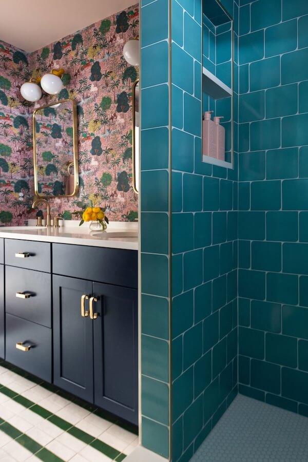 7 Tile Trends Interior Designers Are Loving Right Now - 7 Bathroom with teal tiled shower showcasing the latest tile trends, dark blue vanity with gold handles, floral wallpaper, round mirrors, and globe light fixtures.