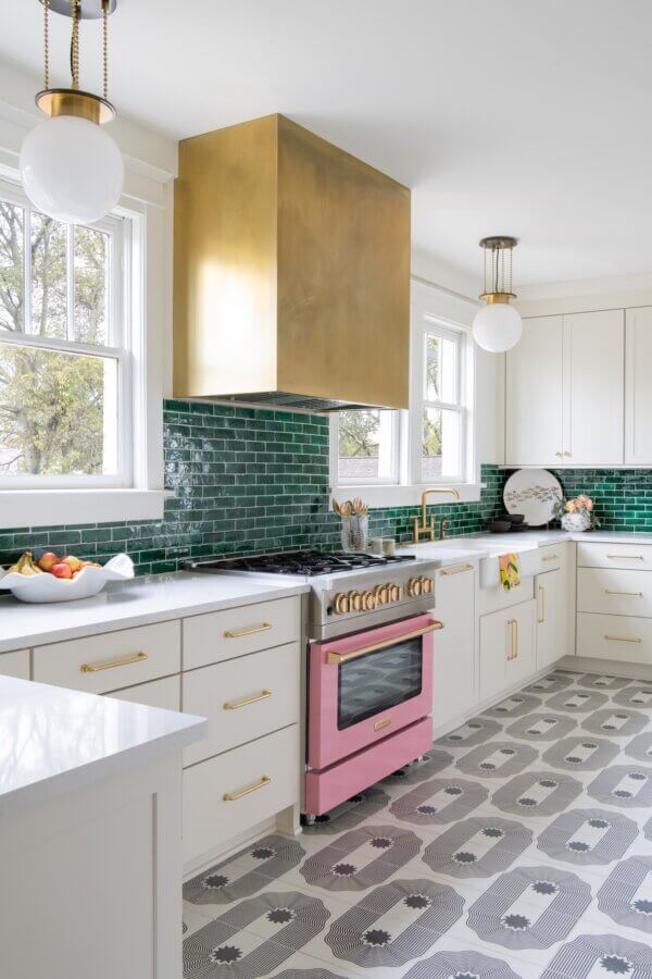 7 Tile Trends Interior Designers Are Loving Right Now - 9 Modern kitchen with white cabinets, green tiled backsplash showcasing the latest tile trends, pink stove, large brass range hood, patterned floor tiles, and two globe pendant lights.
