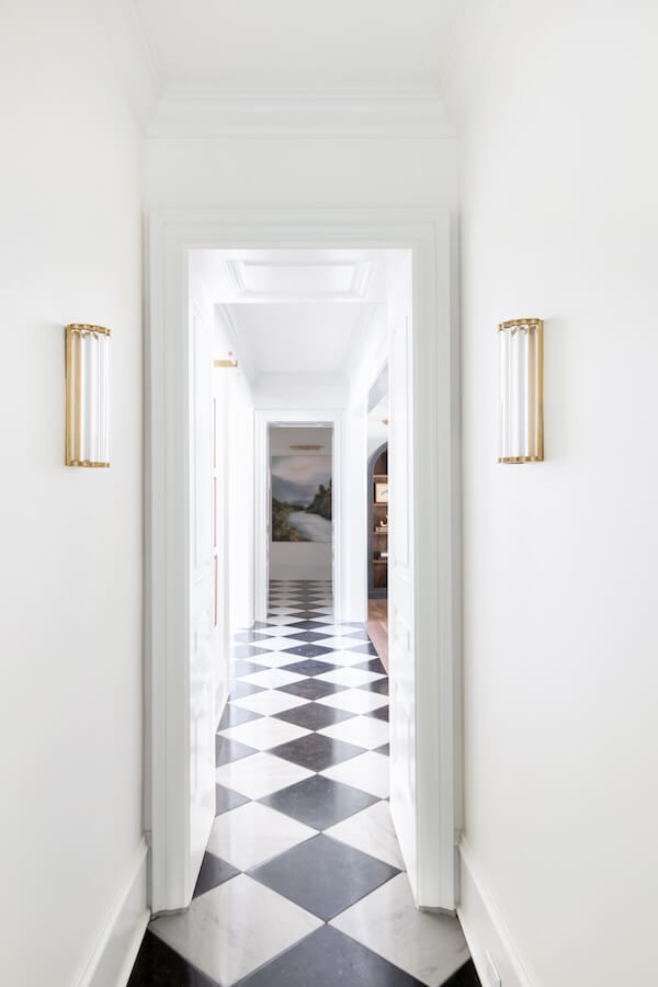 7 Tile Trends Interior Designers Are Loving Right Now - 16 A hallway with white walls and on-trend black and white checkered floor tiles showcases the latest tile trends, illuminated by two wall sconces, leading to a room where a landscape painting is visible at the end.
