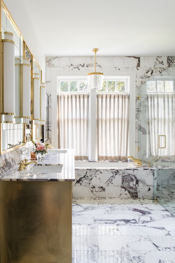 7 Tile Trends Interior Designers Are Loving Right Now - 18 Luxurious bathroom with marble walls and floor inspired by the latest tile trends, gold fixtures, double sink, glass shower, bathtub, and a large window with sheer curtains.