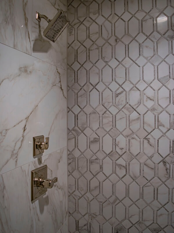 7 Tile Trends Interior Designers Are Loving Right Now - 11 Close-up of a shower with a square metal showerhead, two control knobs, and walls featuring large marble tiles and geometric-patterned tiles, reflecting the latest tile trends.