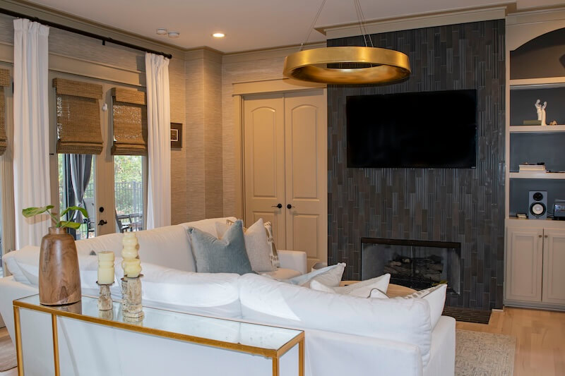 7 Tile Trends Interior Designers Are Loving Right Now - 22 Modern living room with white sofas, a glass-top console table, a black tile fireplace showcasing the latest tile trends, wall-mounted TV, and gold circular ceiling light. Built-in shelves and windows with shades are visible.