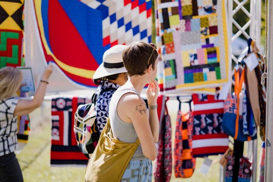 The Best Festivals in the South - 19 Two people admire colorful quilts at an outdoor market or craft fair—an experience found at some of the best festivals in the South—while another person browses in the background.