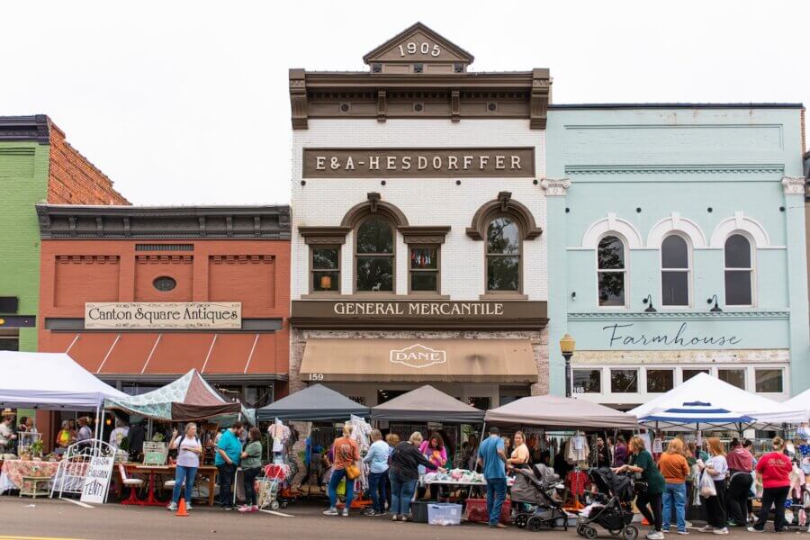 The Best Festivals in the South - 10 Crowds browse vendor stalls at an outdoor market in front of vintage storefronts, including antique and mercantile shops, on a historic Main Street—an atmosphere reminiscent of the best festivals in the South.