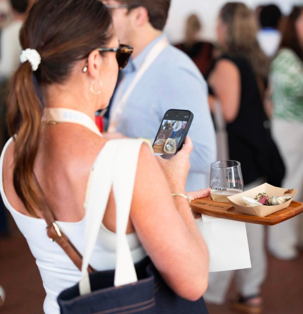 A woman takes a photo of food with her phone while holding a wooden tray with appetizers and a glass at a crowded indoor event, capturing the lively atmosphere reminiscent of the best festivals in the South.