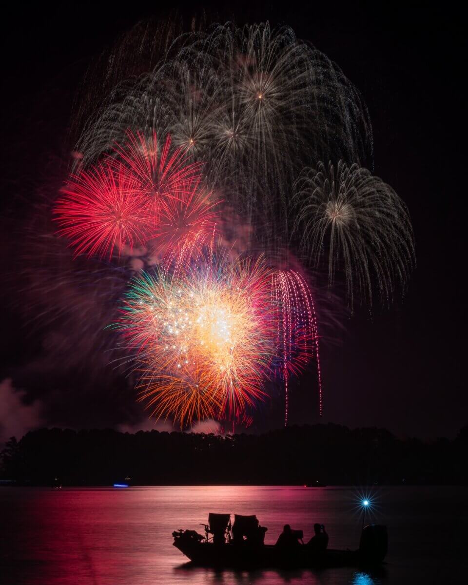 The Best Festivals in the South - 16 Colorful fireworks burst in the night sky over a calm lake, as the silhouette of a boat drifts by and people watch from the water—capturing the magic found at the best festivals in the South.