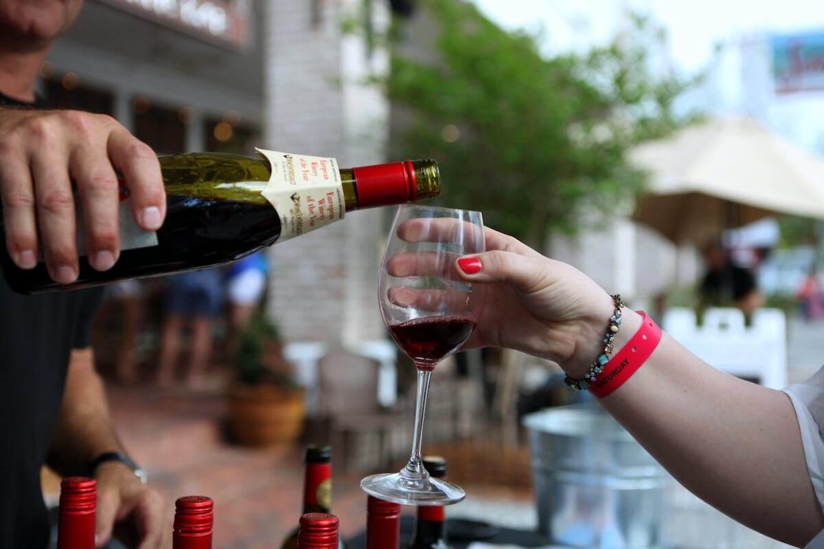 A person pours red wine from a bottle into a glass held by another at an outdoor event, capturing the spirit of Southern festivals, with bottles and a blurred background setting the festive scene.