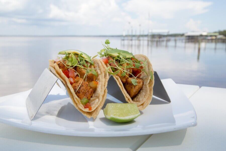 The Best Festivals in the South - 7 Two fish tacos with avocado, diced tomatoes, and microgreens on corn tortillas, served on a plate with a lime wedge—perfect for enjoying outdoors by the water at one of the Best Festivals in the South.