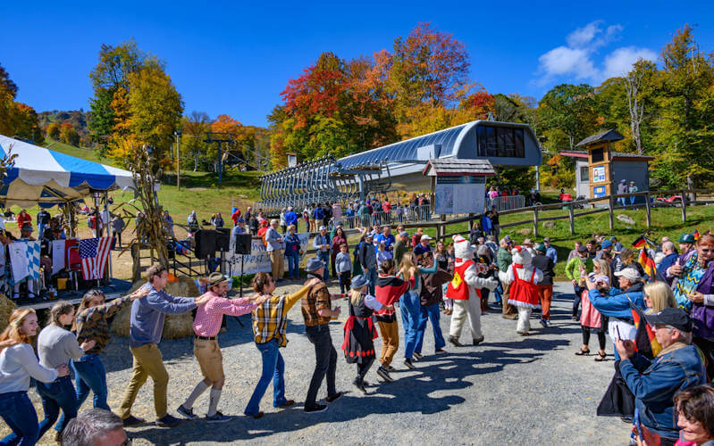 The Best Festivals in the South - 21 People participate in a line dance at an outdoor festival in the South, with autumn foliage, vendor tents, and a ski lift station in the background—one of the Best Festivals to experience in 2026.