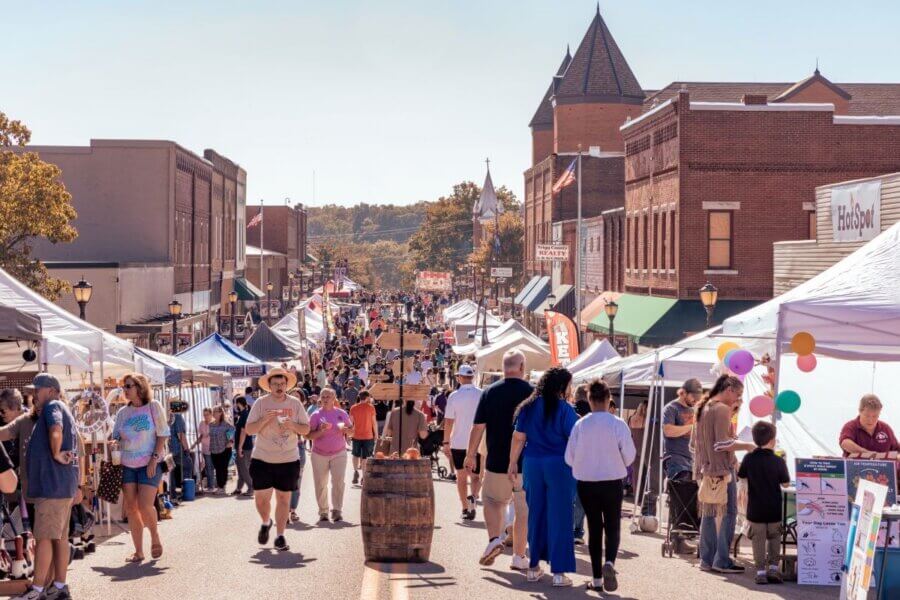 The Best Festivals in the South - 20 A busy street fair, named one of the Best Festivals in the South for 2026, with people walking among vendor tents on both sides; brick buildings line the street under a clear sky.