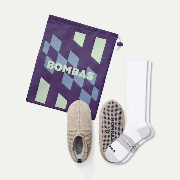 12 In-Flight Plane Accessories You Need - 8 A pair of beige Bombas slippers, white Bombas socks, and a purple drawstring bag with a geometric design and "BOMBAS" text—perfect plane accessories for your next journey.
