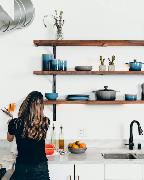 The Tidiness Double Standard - 2 A person with long hair stands at a tidy kitchen counter under wooden shelves holding dishes, jars, plants, and a pot. A bowl of fruit and bottles are neatly arranged on the marble countertop, adding to the sense of tidiness.