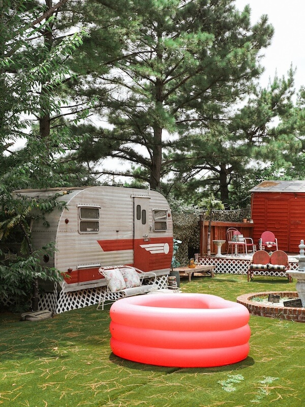 Lil' Graceland: An Elvis-Inspired Retreat Just Minutes from the Real Deal - 4 A small vintage trailer and red chairs sit in a grassy yard with tall trees at Lil Graceland; a pink inflatable pool is in the foreground.