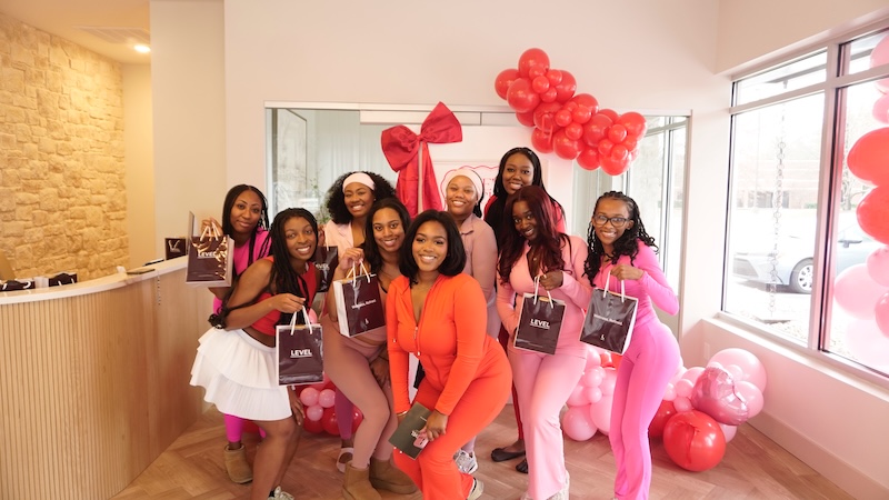 Inside Lea Bryant’s She’s Social Club - 3 Nine women from She’s Social Club, including Lea Bryant, pose indoors with gift bags, standing before pink and red balloon decorations and a large bow. Most are dressed in shades of pink and red.
