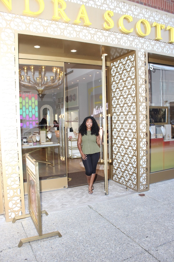 Inside Lea Bryant’s She’s Social Club - 5 Lea Bryant stands smiling in the open doorway of a Kendra Scott store, with jewelry displays visible inside and a decorative gold storefront, welcoming She's Social Club members.