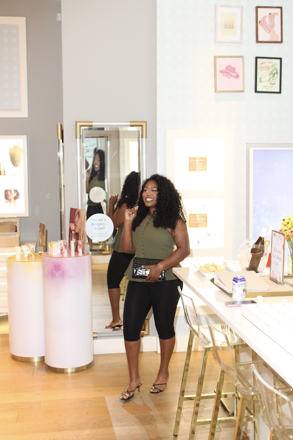 Inside Lea Bryant’s She’s Social Club - 6 Lea Bryant stands smiling in a green sleeveless top and black pants inside She’s Social Club, a well-lit beauty store, holding a purse with cosmetics and chic decor displayed around her.