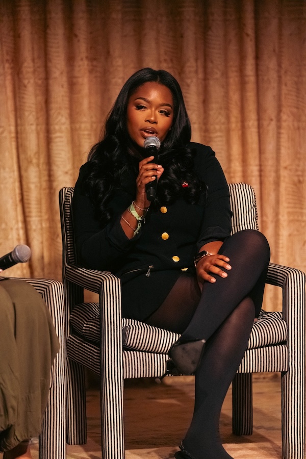 Inside Lea Bryant’s She’s Social Club - 2 Lea Bryant of She's Social Club sits on a striped chair in a black outfit, holding a microphone and speaking, with a curtain in the background.
