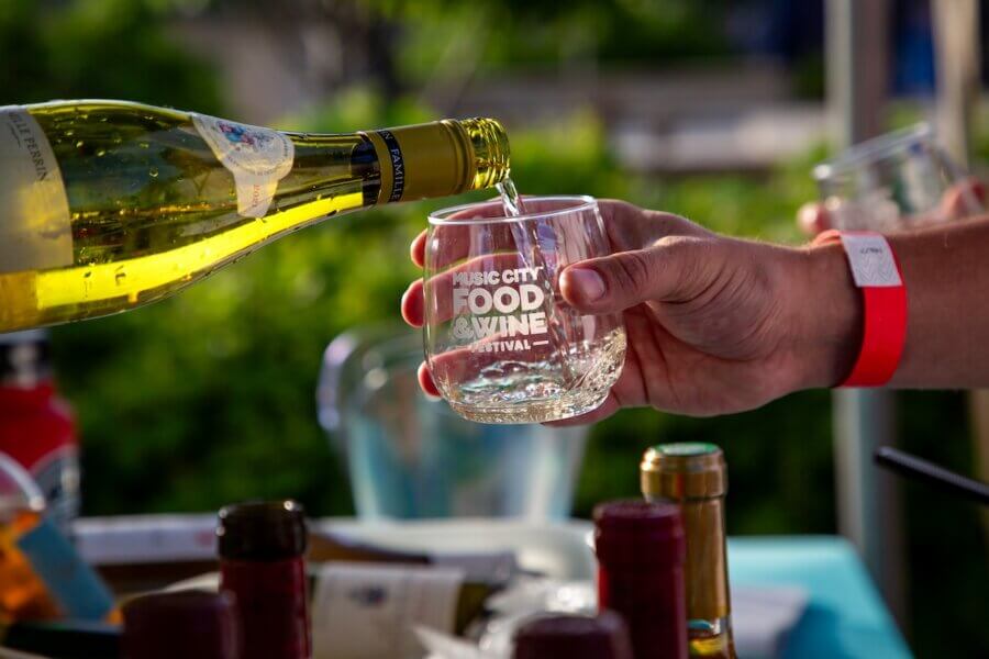 Behind the Scenes with the Director of the Music City Food & Wine Festival - 4 A person pours white wine into a glass labeled "Music City Food & Wine Festival" at an outdoor food festival, with bottles visible on the table.