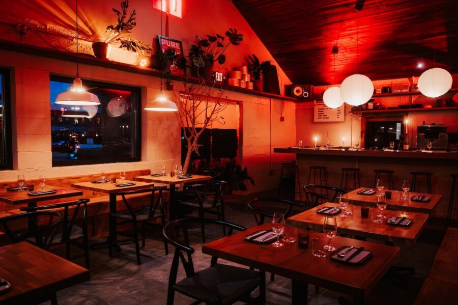 A cozy restaurant interior with wooden tables, black chairs, pendant lights, paper lanterns, and a warmly lit bar area. Table settings are prepared for guests at one of Nashville's inviting new restaurants.