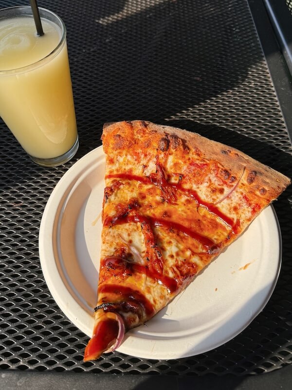 A slice of pizza with barbecue sauce and onions sits on a paper plate next to a yellow drink, enjoyed in the sunlight on a metal mesh table—perfect for sampling what March 2026 brings to new Nashville restaurants.