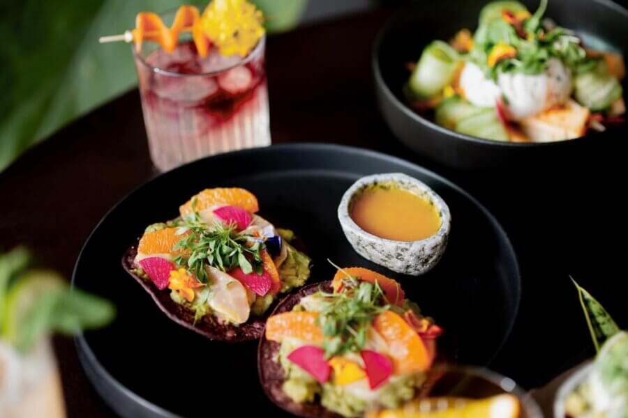 A close-up of a black plate with colorful tostadas topped with guacamole and fresh fruit, a small bowl of yellow sauce, and a cocktail in the background.