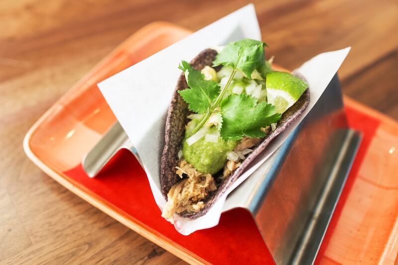 A taco with shredded meat, guacamole, cilantro, and lime in a taco holder on a red tray.