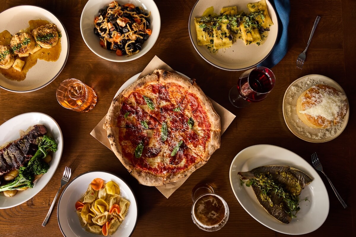 A table set with a pizza in the center, surrounded by various pasta dishes, steak with vegetables, stuffed fish, drinks, and dessert—perfect for sampling the flavors of new restaurants opening in Nashville in March 2026.