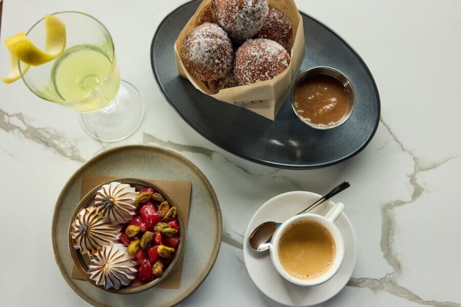 A table set with sugared doughnuts, caramel dipping sauce, a lemon cocktail, a dessert topped with meringue and berries, and a cup of espresso hints at the indulgent offerings from new Nashville restaurants in March 2026.
