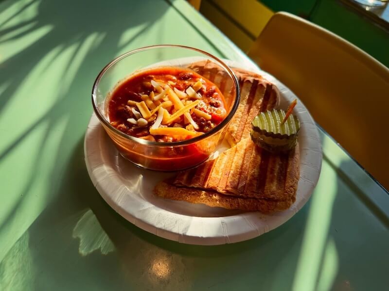 A bowl of chili topped with shredded cheese, served with grilled bread slices and pickle chips on a paper plate.
