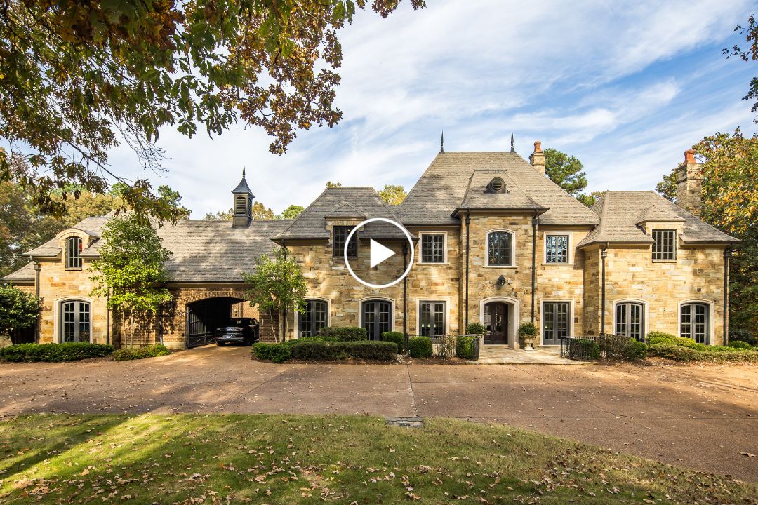 Elegant stone home at 214 Mountain Brook Cv in Eads TN, featuring multiple chimneys, arched windows, and a covered driveway entrance, set among trees and a leaf-covered lawn.
