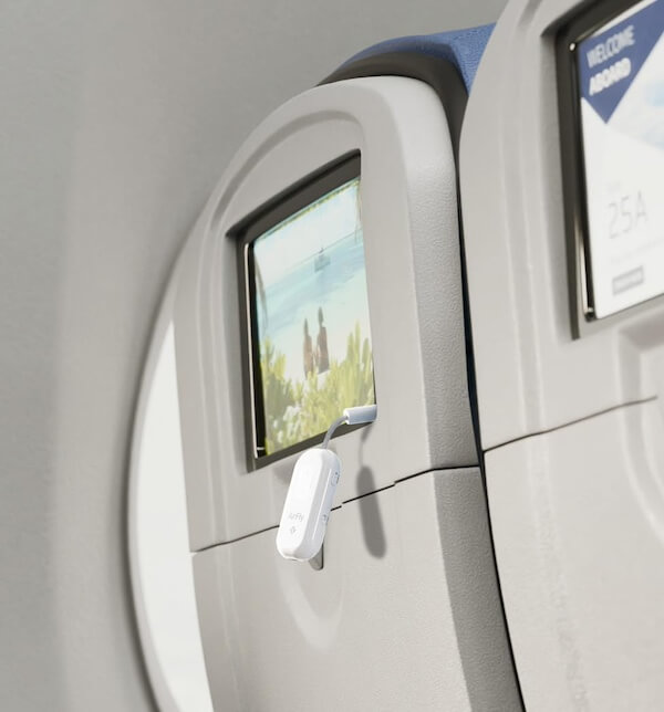 12 In-Flight Plane Accessories You Need - 7 Close-up of an airplane seatback screen displaying a beach scene, with a small white electronic device—one of the must-have plane accessories—plugged into a USB port below the screen.